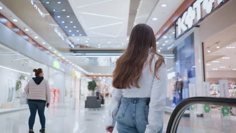 dama con teléfono en el bolsillo trasero llevando una bolsa de compras baja de la escalera mecánica y se vuelve en un centro comercial moderno, el cabello largo fluye mientras se mueve a través de un espacio comercial bien iluminado con compradores ocupados en el fondo