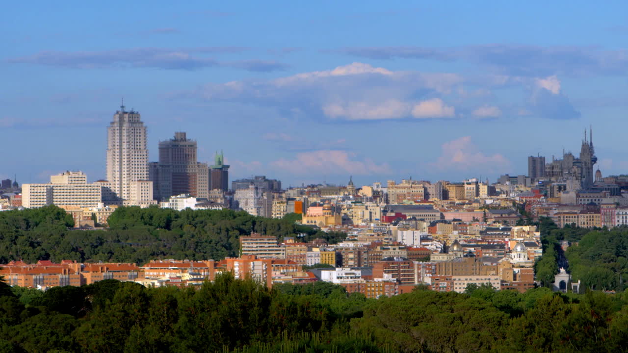 Madrid’s skyline rises through rich layers of trees and rooftops, beneath a wide sky filled with billowing white clouds.