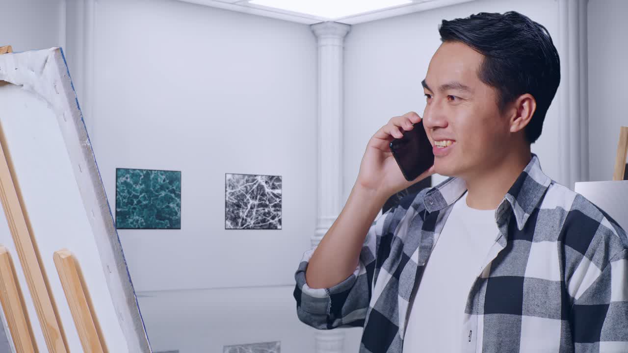 갤러리에서 오일 페인트와 으로 캔버스에 그림을 그리는 동안 스마트폰으로 이야기하는 아시아 남성 예술가의 클로즈업