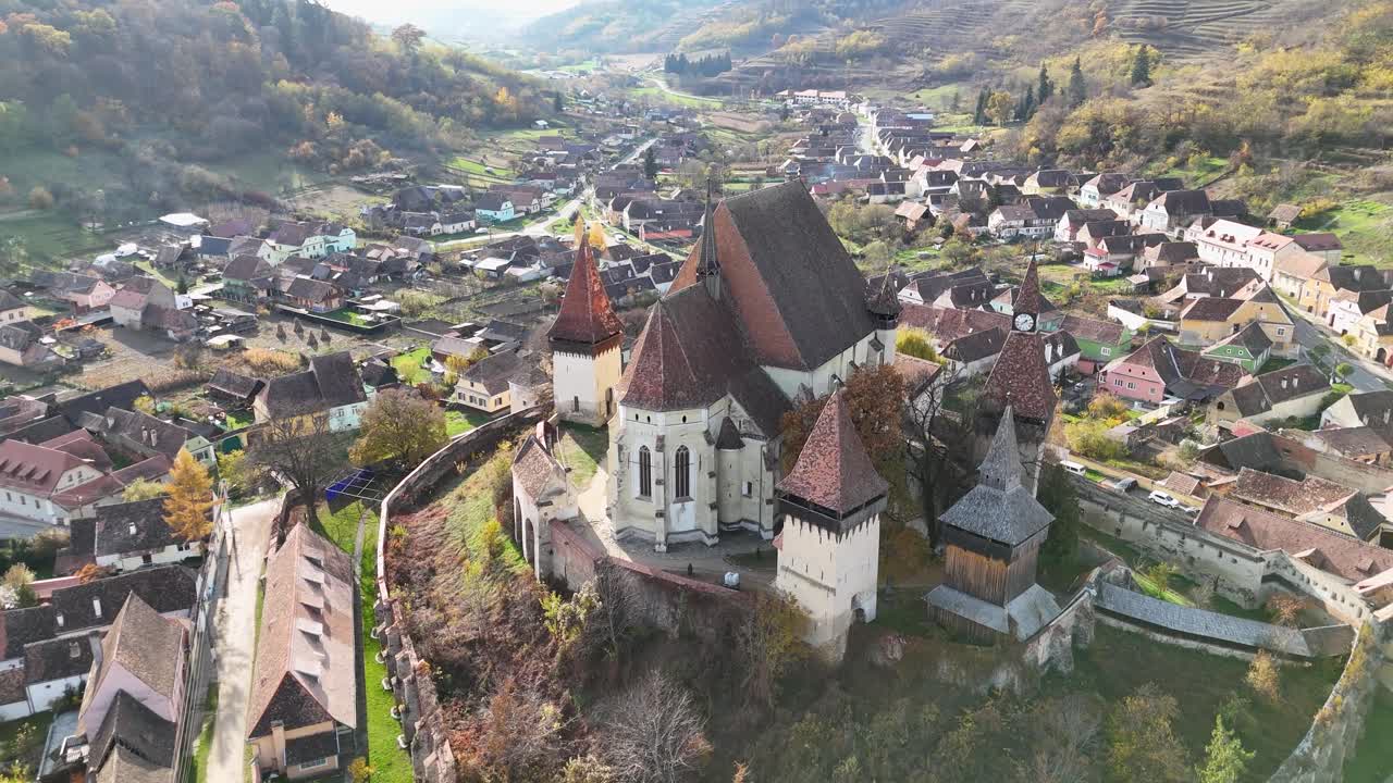The camera soars above the church, showcasing its impressive architecture and fortified walls, while offering a panoramic view of the picturesque village below.