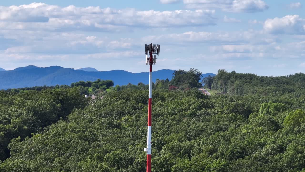 Tall red and white communication tower rises above dense green forest with residential buildings and mountains in background. Aerial view in Suburb of American town in summertime. Wide shot