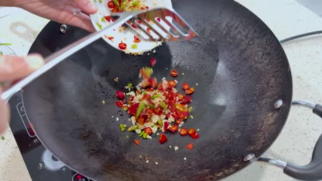 a hand pours chopped red chili and minced garlic into a preheated wok on a stovetop, under bright kitchen lighting, for spicy cooking preparation