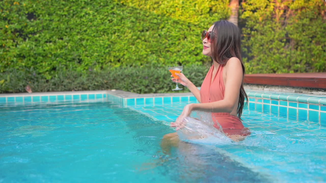 Beautiful girl in glasses and swimsuit sitting on the pool, drinking cocktail and throwing water