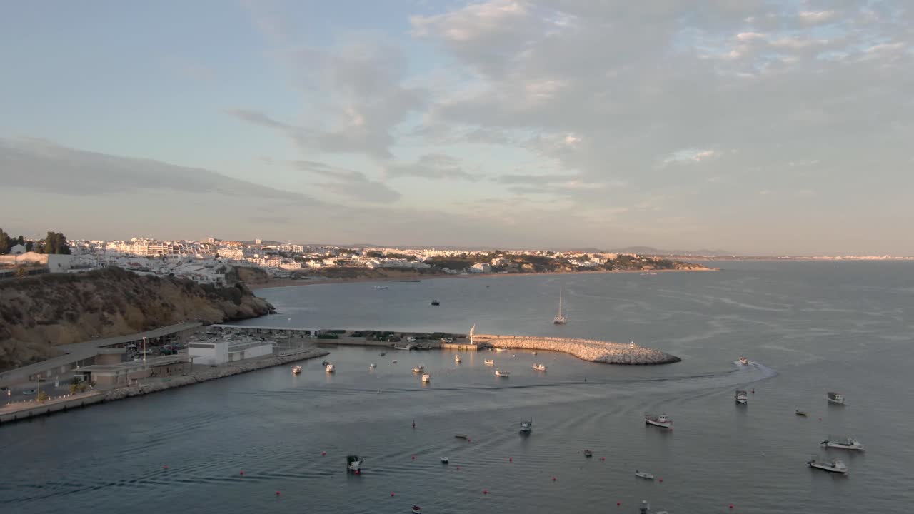 Aerial Static Shot of Boast leaving Fishing Harbor during sunset with cloudy sky. Beach and town in the distance.