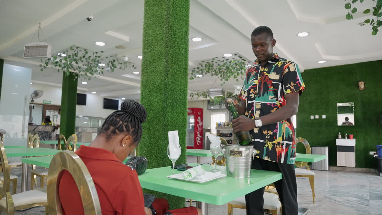 Coppia afroamericana a un tavolo verde del caffè, uomo che apre lo champagne e prepara un brindisi romantico, donna in abito rosso seduta, arredamento moderno con piante, illuminazione soffusa, conversazione intima, cameriere nelle vicinanze, atmosfera celebrativa.