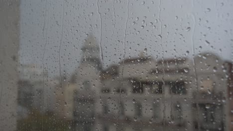 a slow-motion close-up view of heavy raindrops landing on a window glass, with an urban city landscape in the background