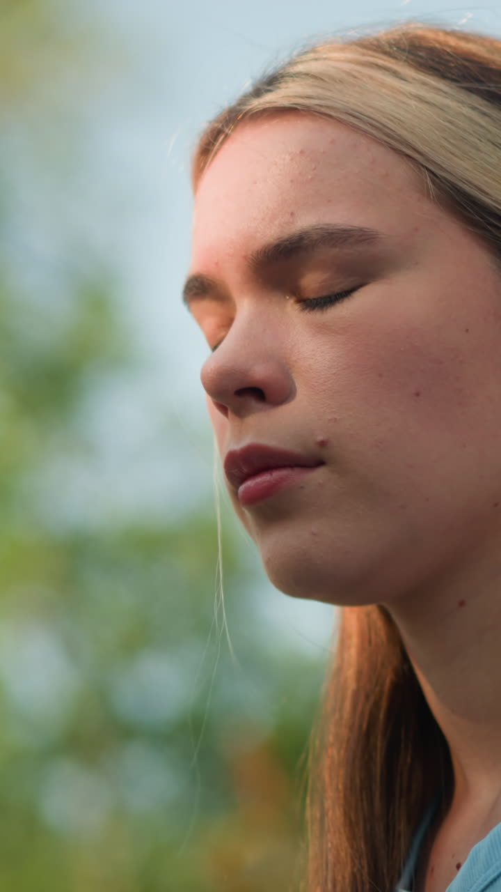 mujer joven al aire libre, tocando suavemente su cabello mientras la luz del sol ilumina suavmente su cara, con una camiseta cian, con el cabello fijado hacia atrás con una aguja negra, bajo un cielo azul, rodeada de vegetación borrosa