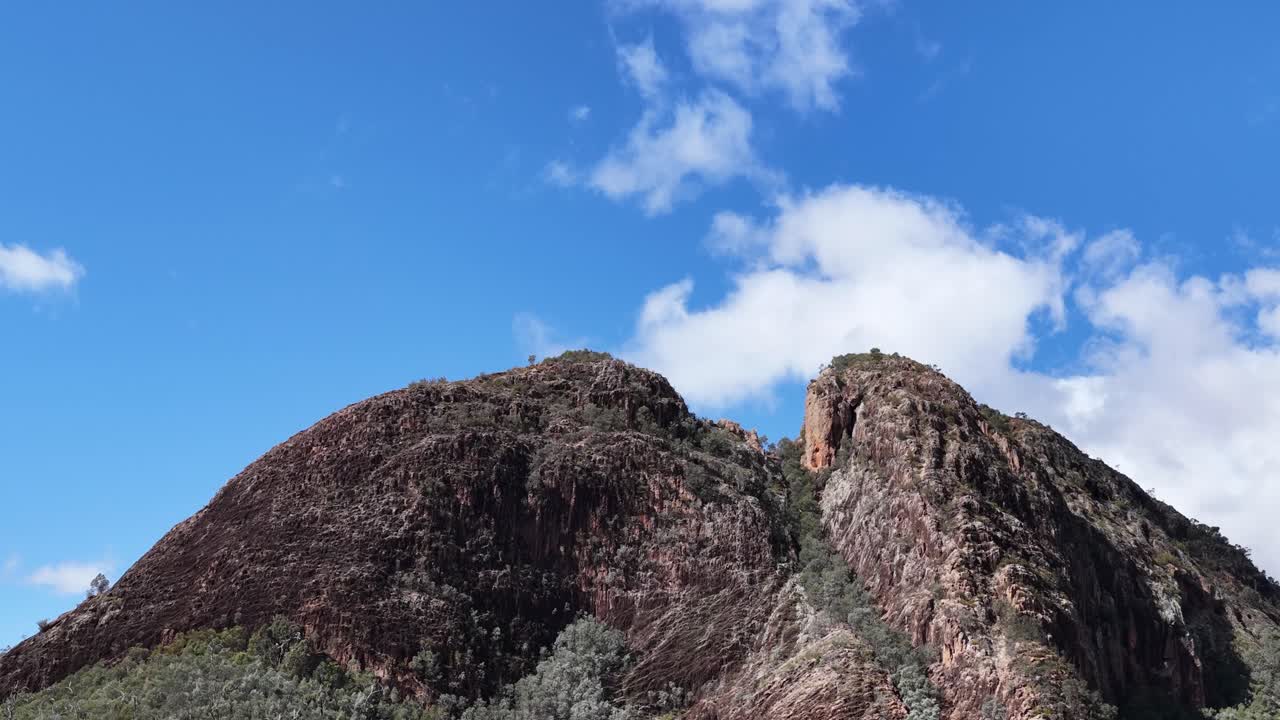 Camera slowly pans across Split Rock’s rugged summit under bright daylight, revealing rocky textures, sparse vegetation, and a vivid blue sky with scattered clouds