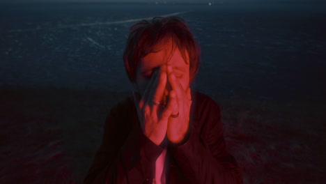 man covering his face in red light beside frozen lake at night