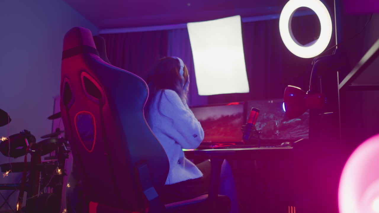 back view of excited gamer raising hands in celebration surrounded by glowing ring light, monitors, red condenser mic, drum set, and studio gear, immersed in vibrant streaming