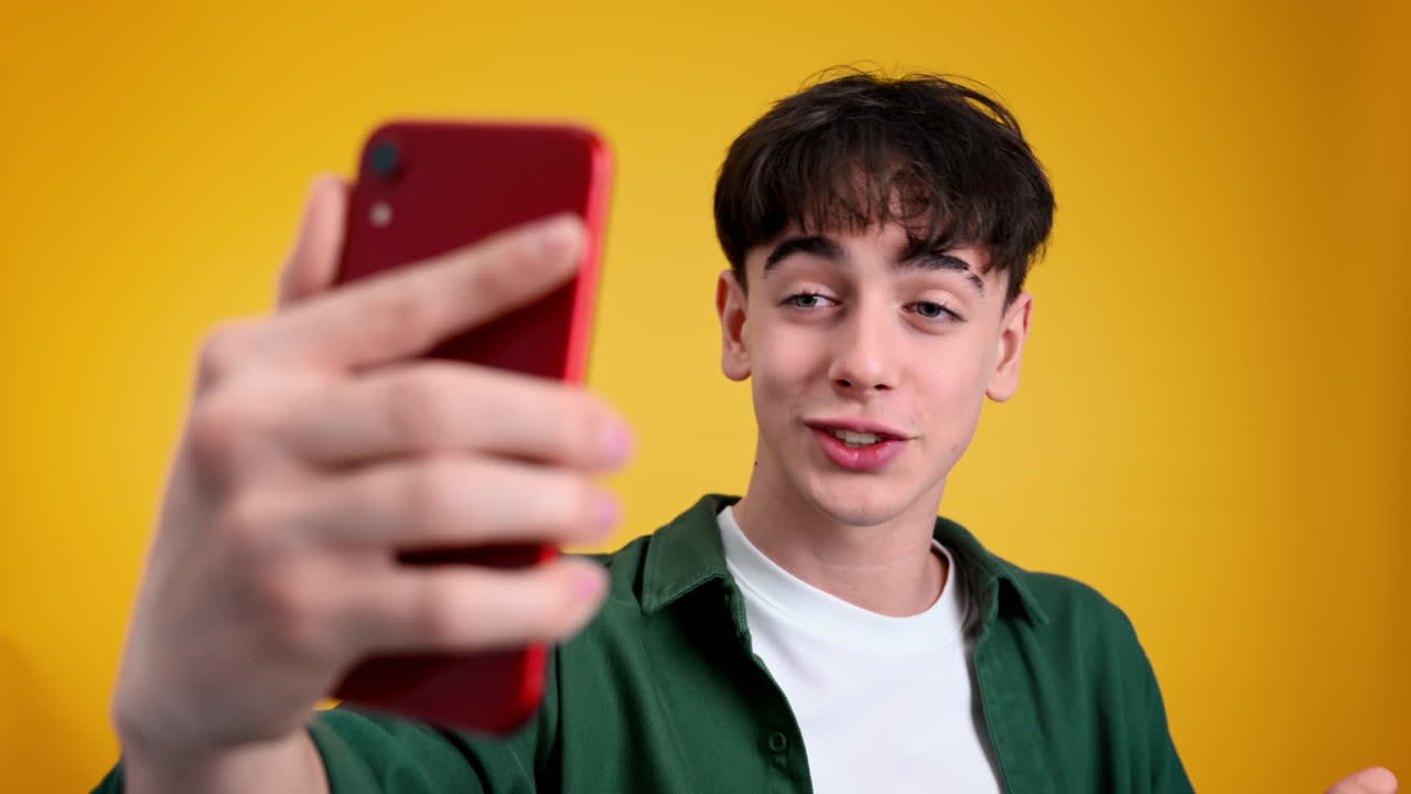 Young man talking while filming himself on the phone and gesturing on a yellow background