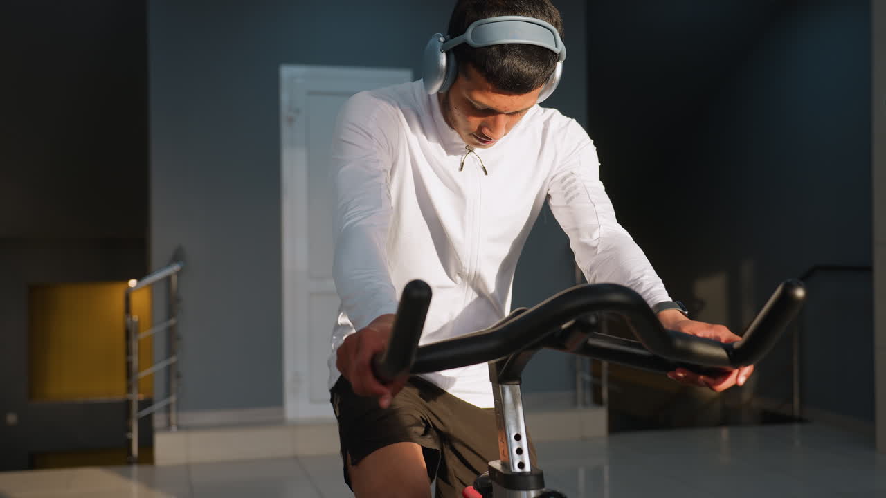 Young boy in white sportswear walks to stationary bike and climbs on to begin cycling under sunlight filtering through gym windows, with white door and equipment visible in blurred background