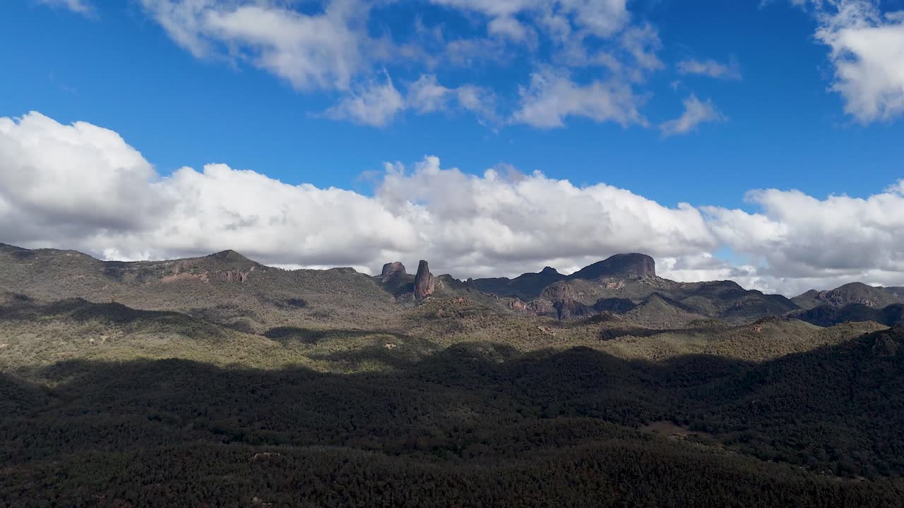 Drone camera smoothly pans across rugged volcanic mountain peaks under a partly cloudy sky in Warrumbungle National Park, with dramatic midday lighting