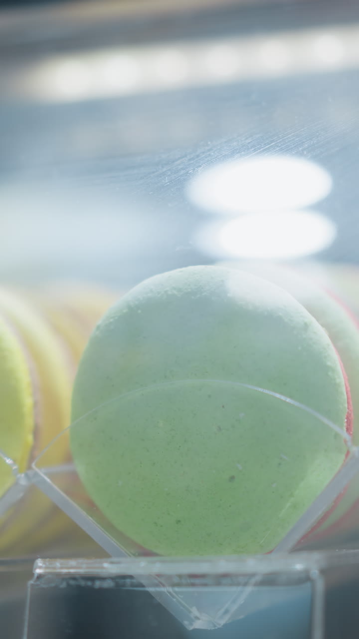 primer plano de varios macarones coloridos dispuestos en una vitrina con luz que se refleja en el vidrio, mostrando macarones amarillos, verdes y blancos