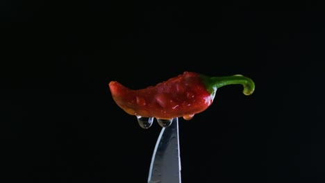 red wet jalapeno pepper on a black background pinched from bottom by a silver knife with shots of flashing light coming from lower left side