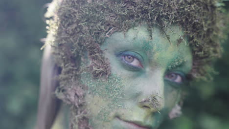 Close-up-of-a-female-avatar-wearing-green-bodypaint-looking-towards-the-camera-in-a-forest,-in-the-style-of-nature-inspired-forms