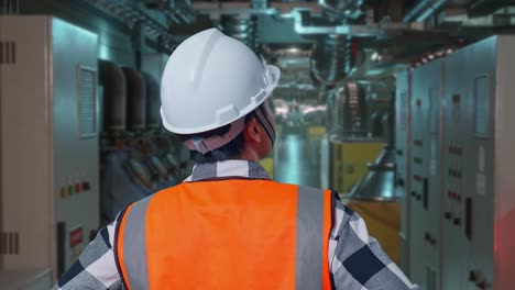 close up back view of a male engineer wearing safety helmet looking around while standing with arms akimbo in engine control room, work of electrical generators