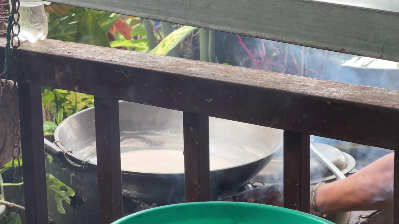Boiling pot of coconut milk behind a wooden railing in an outdoor kitchen. Great for rural cooking, cultural food scenes, or authentic culinary storytelling