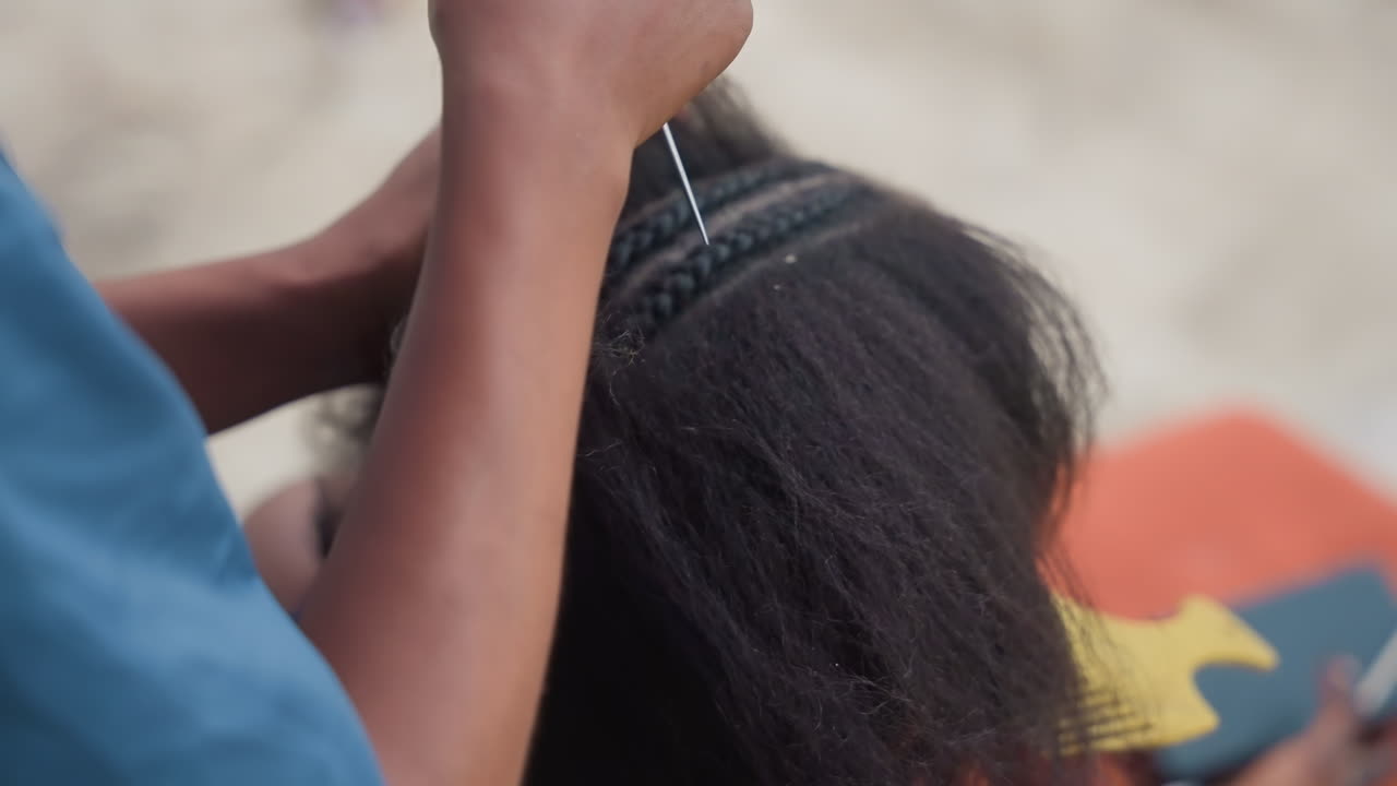 Primo piano di capelli neri pettinati all'aperto. Stilista concentrato che usa un pettine a forchetta di metallo per sezionare e districare. Mani ritmiche che intrecciano una treccia, ambientazione su una panchina urbana. Separazione precisa e tecnica pulita, attenzione ravvicinata.