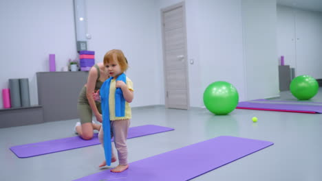 close-up of a little girl wearing a blue cloth around her neck as she plays, while a woman beside her adjusts her outfit in a modern gym setting, with fitness balls visible in the background