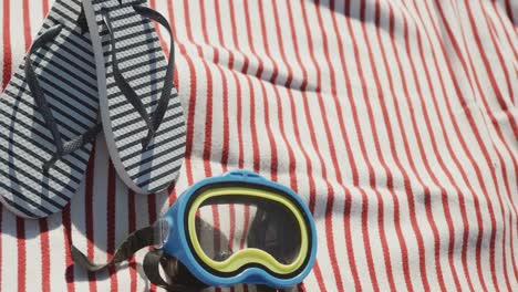 Close-up-of-goggles,-flip-flops-and-towel-on-beach,-in-slow-motion,-with-copy-space