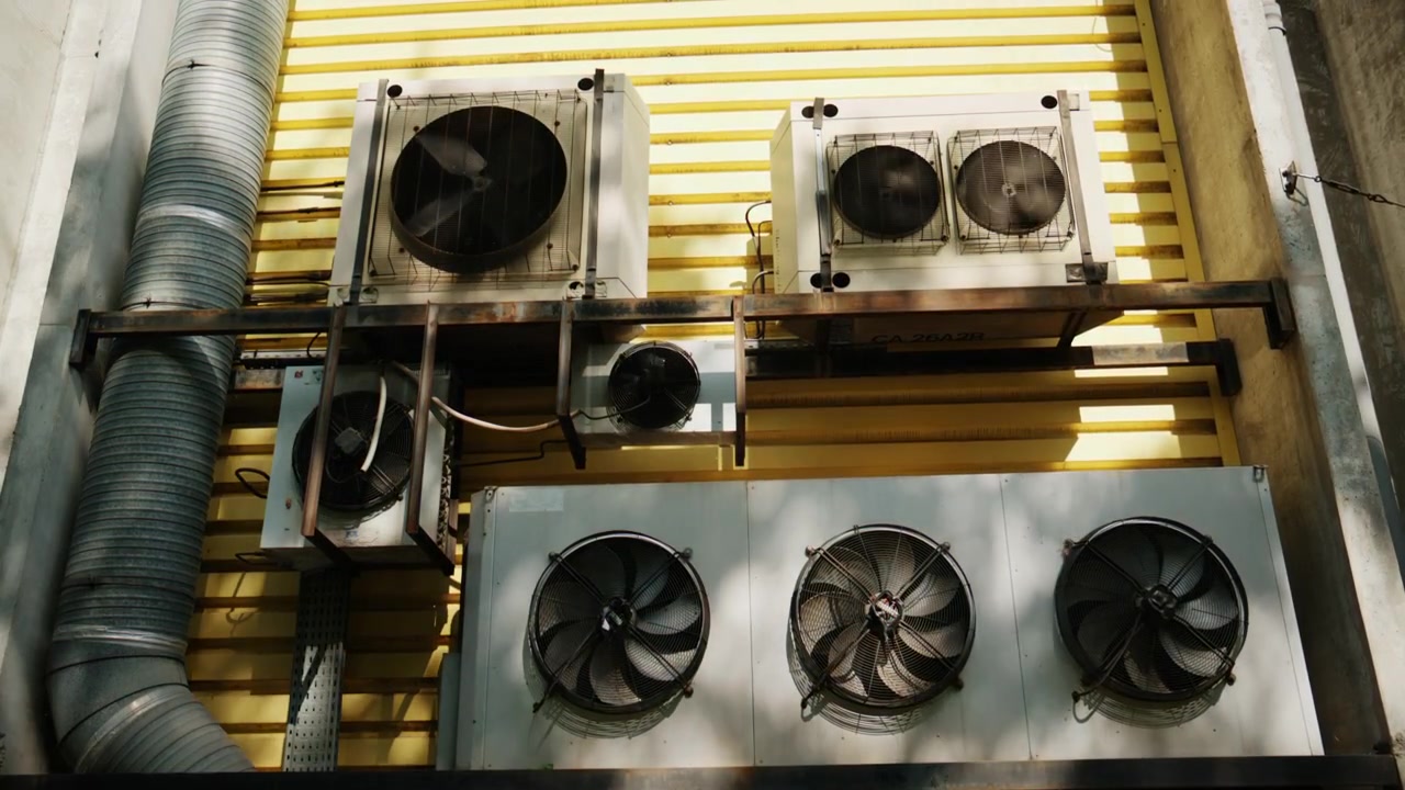 Exterior industrial ventilation and air conditioning units mounted on a yellow metal wall, with large fans, ducts, and sunlight shadows