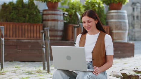 joven feliz usando una computadora portátil teniendo una conversación de chat de llamada de video mientras está sentada en la calle de la ciudad