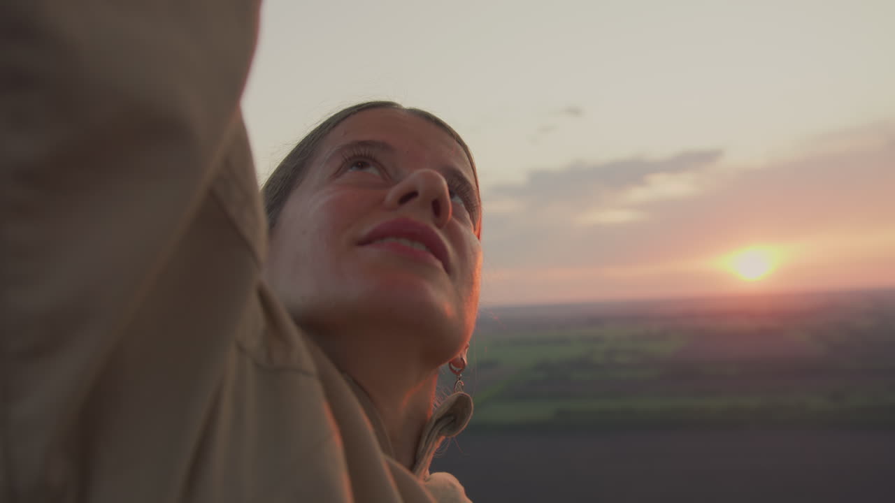 Woman in beige jacket raises arm to adjust burner inside hot air balloon at golden hour, warm light illuminating face as she looks upward with calm determination over open sunset lit landscape