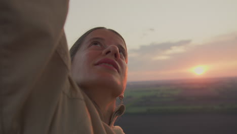woman in beige jacket raises arm to adjust burner inside hot air balloon at golden hour, warm light illuminating face as she looks upward with calm determination over open sunset lit landscape