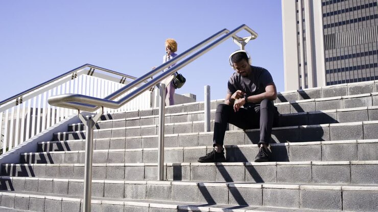 Seated man checking watch, woman in lilac suit appearing walking down urban stairs talking on phone