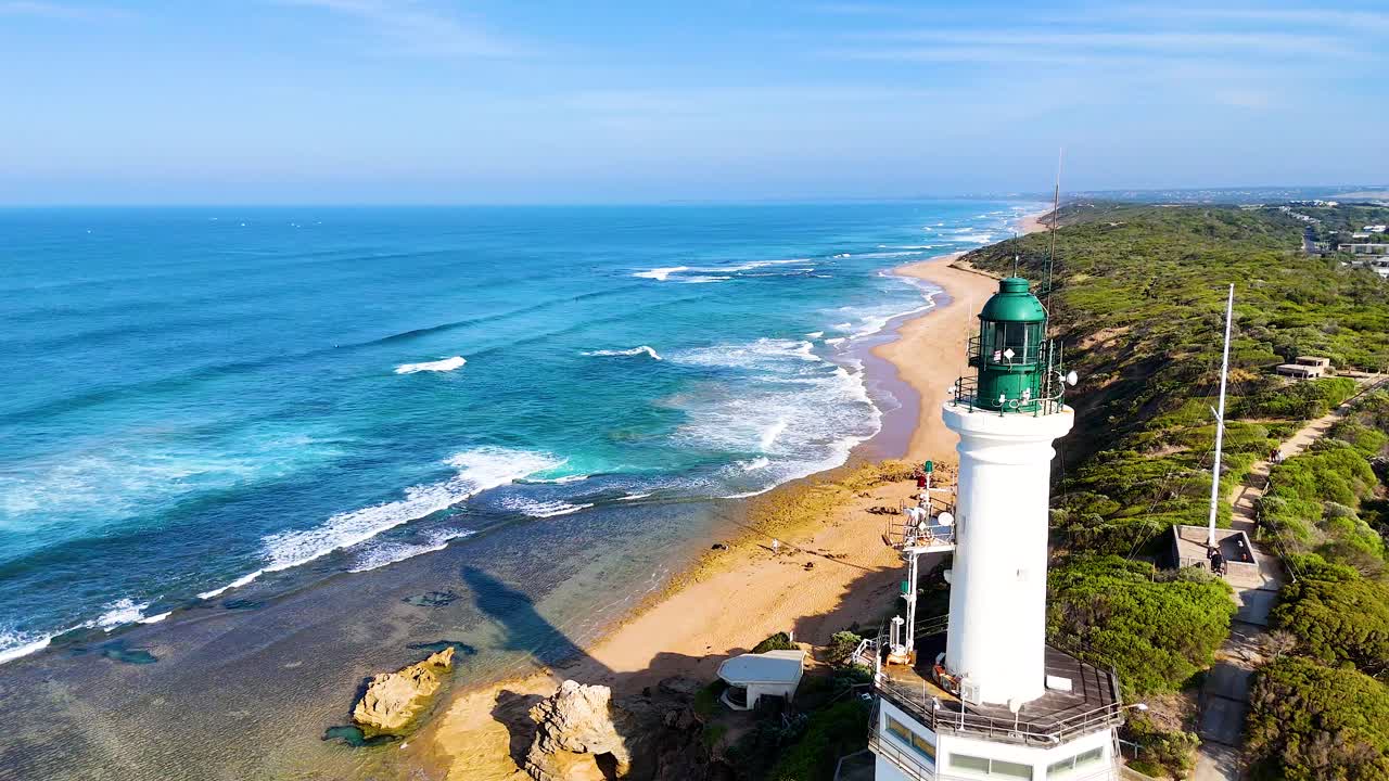 Drone footage captures the scenic Point Lonsdale lighthouse against a backdrop of ocean waves and sandy beaches under clear skies