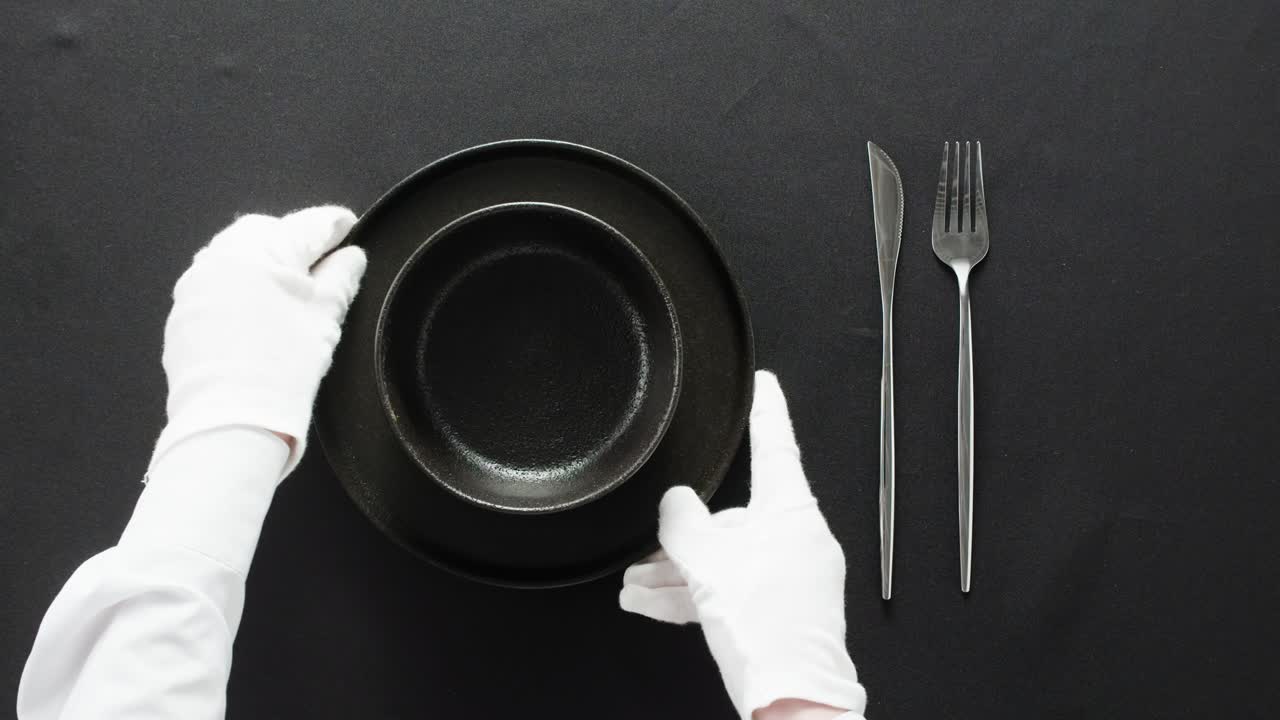 Minimalist table setting with white plate, fork, and knife arranged neatly, and a gloved hand adjusting cutlery.