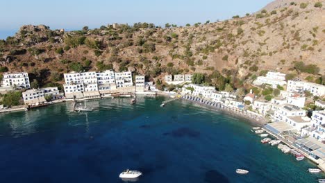 Drone-view-in-Greece-flying-over-blue-sea-in-Loutro-small-white-house-town-and-small-boats-next-to-a-hill-on-a-sunny-day