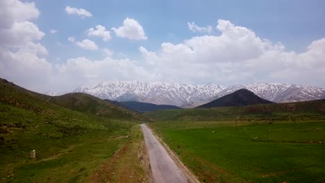 Endlose-Gerade-Asphaltstraße-Inmitten-Einer-Grünen,-Malerischen-Rasenwiese-Mit-Der-Hohen-Schneebergkette-Und-Blauem-Himmel-Mit-Weißen-Wolken-Auf-Der-Reise-Ins-Iranische-Hochland-Am-Sonnigen-Sommertag