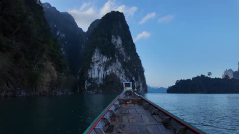 front of traditional wooden boat cruising over calm water in thailand moving towards limestone rock formations and mountains with dense native rainforest