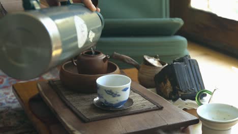 water being poured from a thermos into a clay teapot on a tea table next to other tea ware