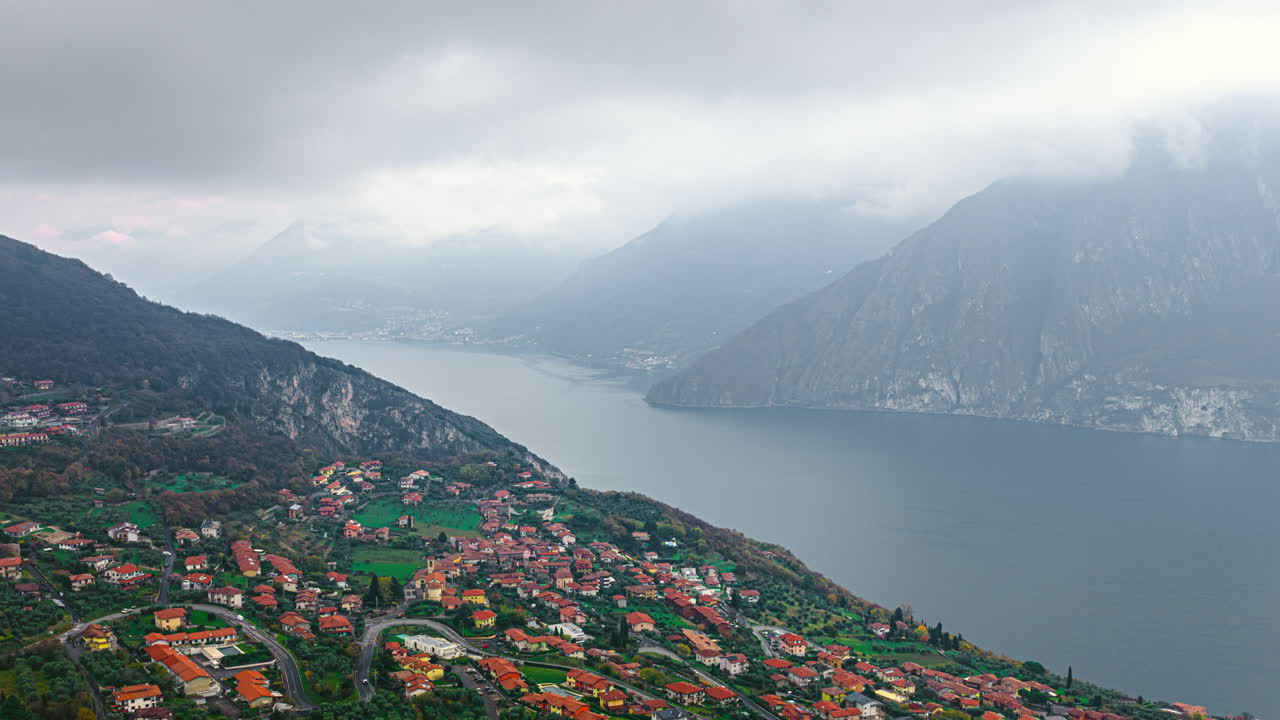 Lago d’Iseo’ya bakan, İtalyan yamaç kasabasının ve yeşil terasların puslu dağlara doğru uzandığı, gölün üzerindeki hareketli bulutları yakalayan kasvetli bir hızlandırılmış çekim.