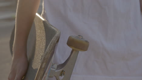 closeup on a skateboard deck, truck and wheel held by a skater wearing a white t shirt at golden hour