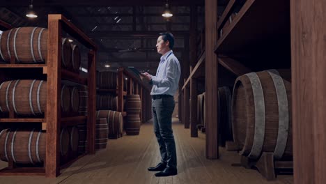 full body side view of asian male professional worker standing with his tablet in cellar of winery, he observes by looking around before he come to concentrating with his tablet