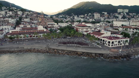 the-boardwalk-of-Puerto-Vallarta-at-dawn,-showcasing-the-city,-church,-mountains,-and-the-famous-arches-of-Puerto-Vallarta-in-the-background,-illuminated-by-the-sun-hitting-the-mountains