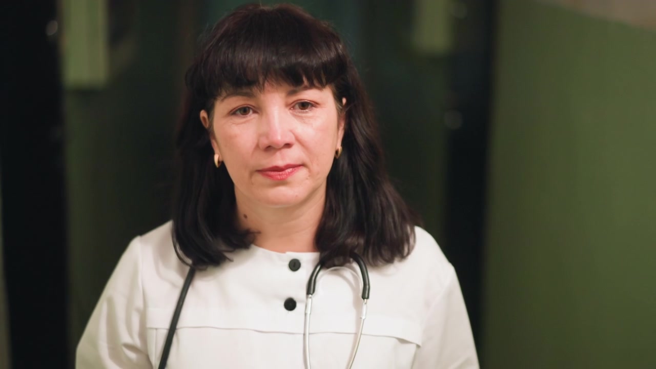 Clinica donna sorridente in un corridoio luminoso, stetoscopio visibile, espressione accogliente, luce naturale che filtra dalla porta, postura attenta al paziente, sottile rassicurazione, scena di controllo di routine, empatica.