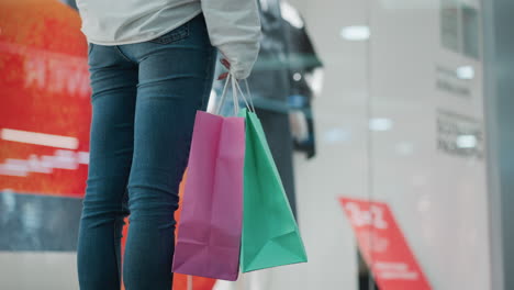 back view of woman standing with shopping bags gently swaying in a well-lit mall, background shows clothing displays in stores with vibrant colors and modern retail environment