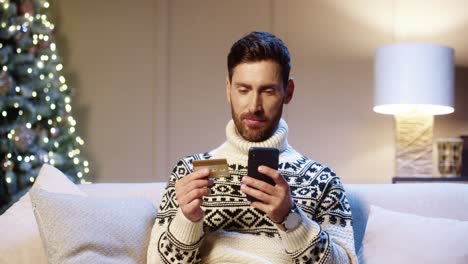 Close-Up-Portrait-Of-Cheerful-Young-Man-Makes-Purchase-On-Internet-Buying-Xmas-Gifts-On-Cellphone-Paying-With-Credit-Card-At-Home-Near-Glowing-Tree