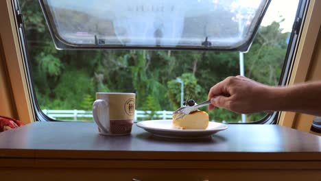 life in camper van. man hand taking bite of cake standing on table with hot tea or coffee near open window with nature view. road trip lifestyle. concept of freedom and vacation.