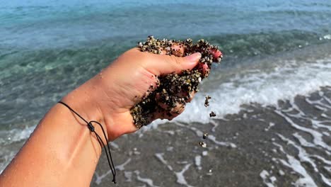 Hand-rubbing-tiny-little-rocks-at-the-beach-with-turquoise-sea-water-and-waves-in-Manilva-Spain,-fun-sunny-summer-day,-4K-shot