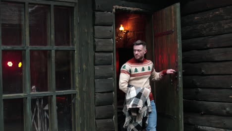 handsome bearded man walks out of the old rural house. fit man in sweater goes somewhere holding warm plaid. attractive man at wooden house background.