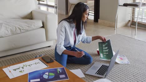 mujer de raza mixta sentada en el suelo en casa usando una computadora portátil y escribiendo en un cuaderno