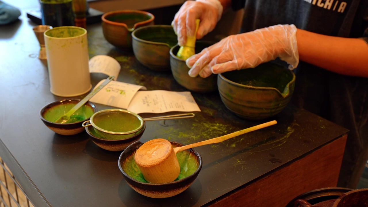 陶器の茶碗と竹製の道具を使った抹茶の準備。緑の粉とふるいがあるカウンタートップに、手袋をはめた手が濃い色の陶器を並べている。