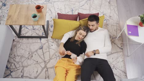 una joven pareja feliz divirtiéndose, riendo, jugando juegos usando un teléfono inteligente en casa.