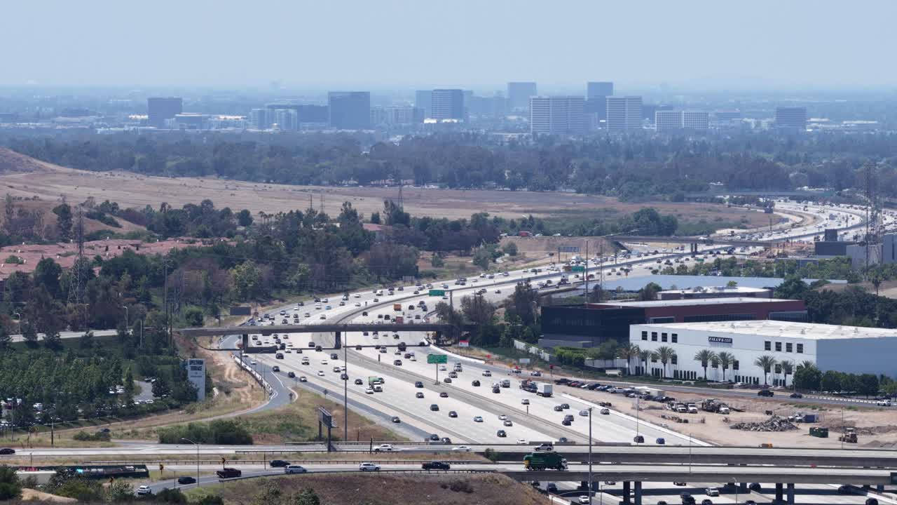 Aerial drone footage over the 405 Freeway in Irvine, California. The shot captures busy highway traffic with the business center skyline of Orange County in the background under sunny skies
